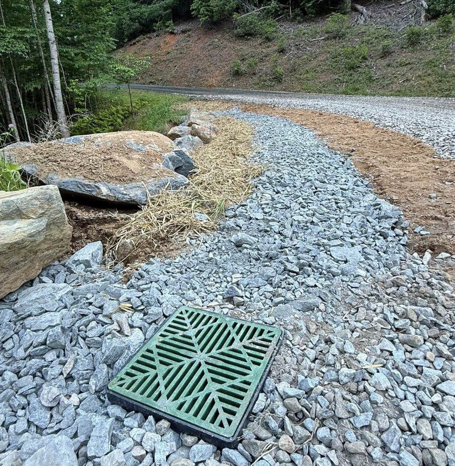 A catch basin set in riprap alongside a gravel driveway to carry water away.